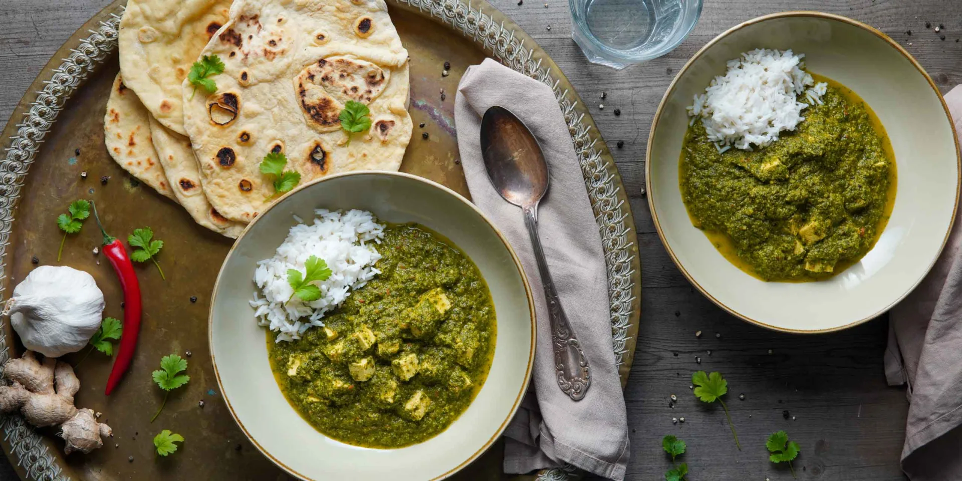 Vegan palak paneer in two bowls with naan bread and water, seen from above. Decorated with fresh ingredients like chili and garlic.