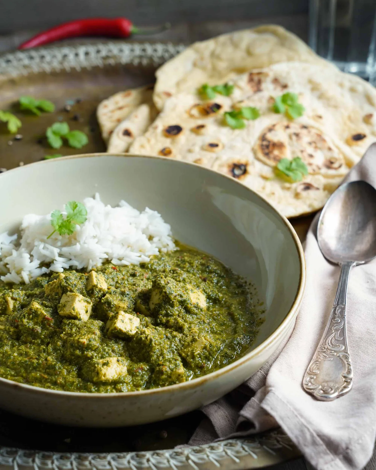 Vegan palak paneer in two bowls with naan bread and water, close-up from the side. Decorated with fresh ingredients like chili and garlic.