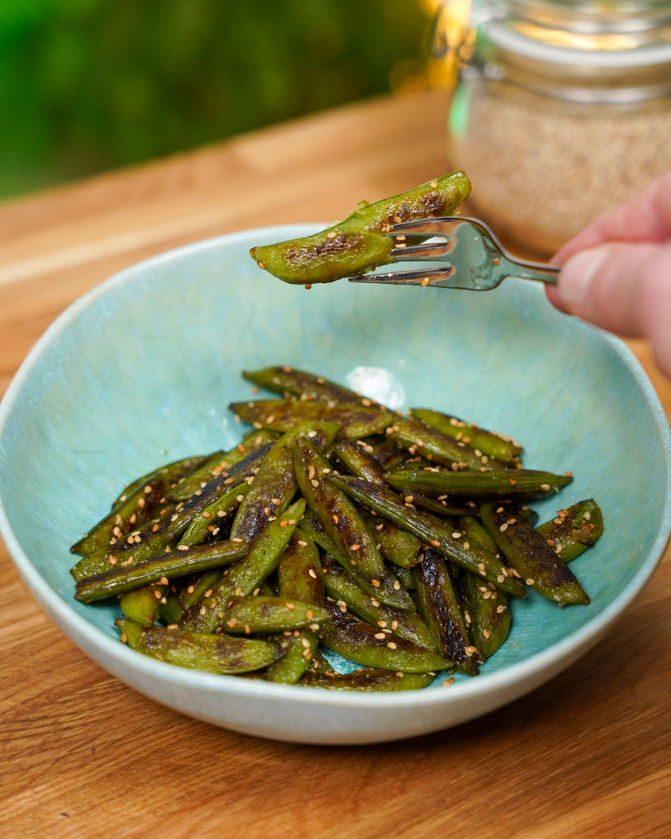 Zuckererbsenschoten in einem Teller angerichtet. Eine Schote mit einer Gabel aufgepickt und in die Kamera gehalten.