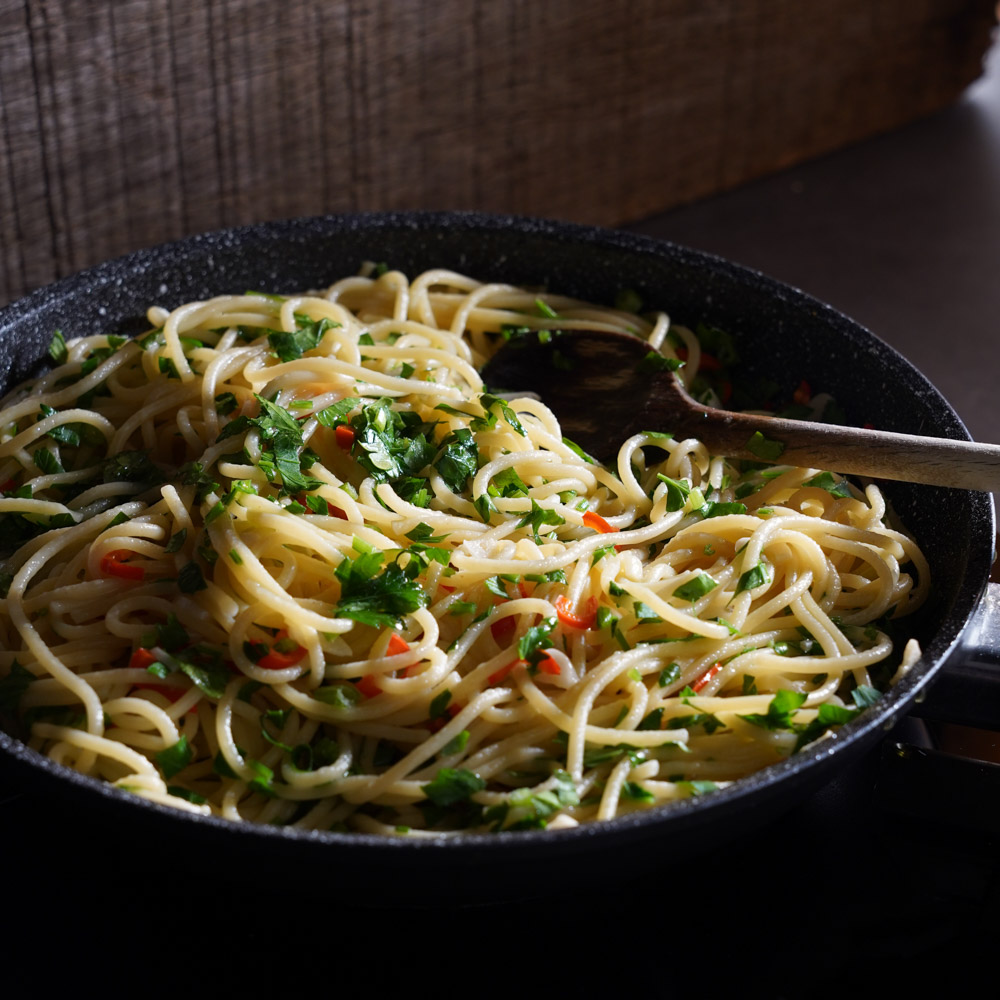 Detailaufnahme von Chili. Im Vordergrund vegane Spaghetti Aglio e olio Spaghetti zieht in der Pfanne restliches Nudelwasser und Aromen auf. Mit Petersilie verfeinert.