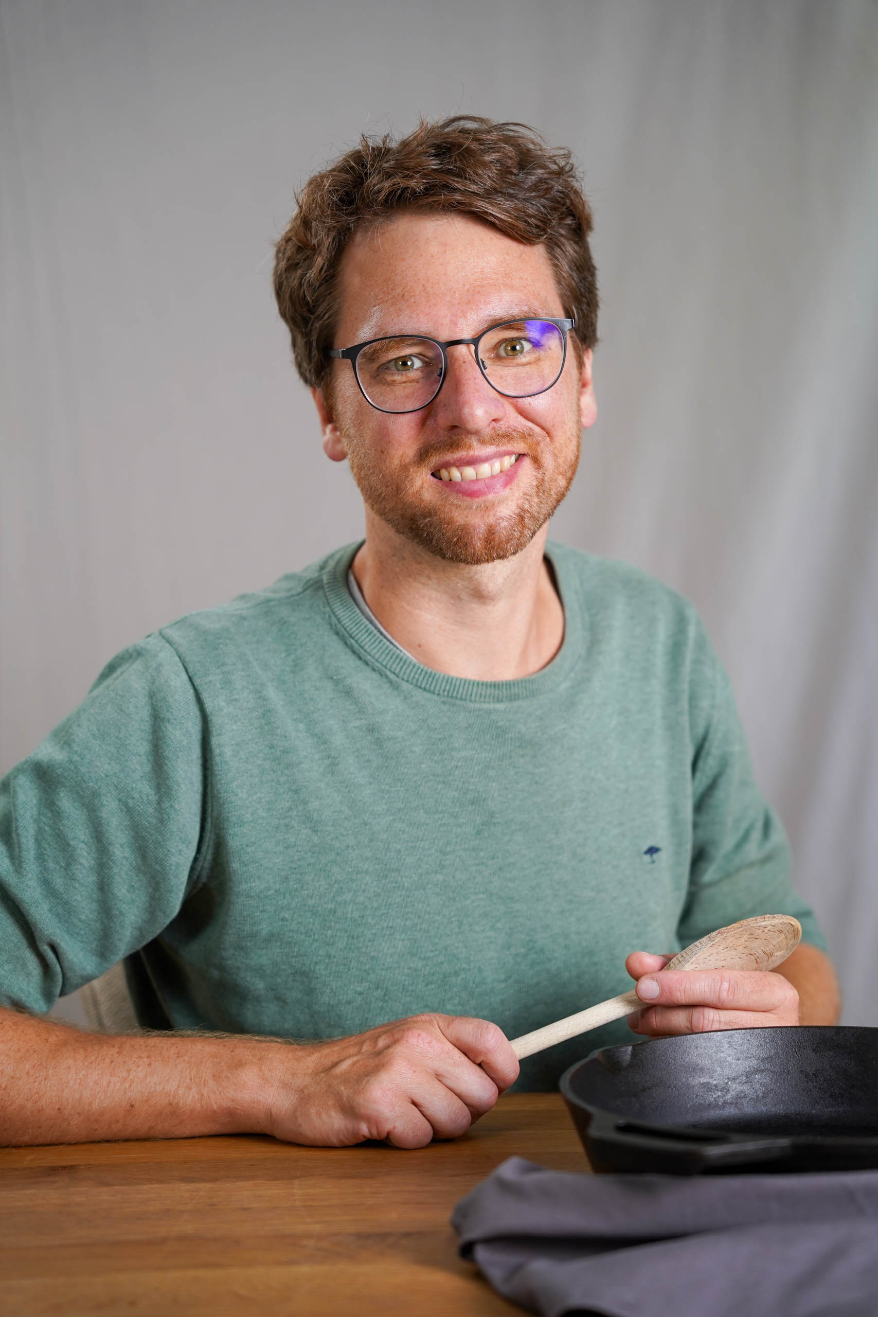 Portrait Ueber mich easy peasy vegan2 Portrait, wo ich an einem Holztisch sitze. Kochlöffel in der Hand und gußeiserne Pfanne vor mir.